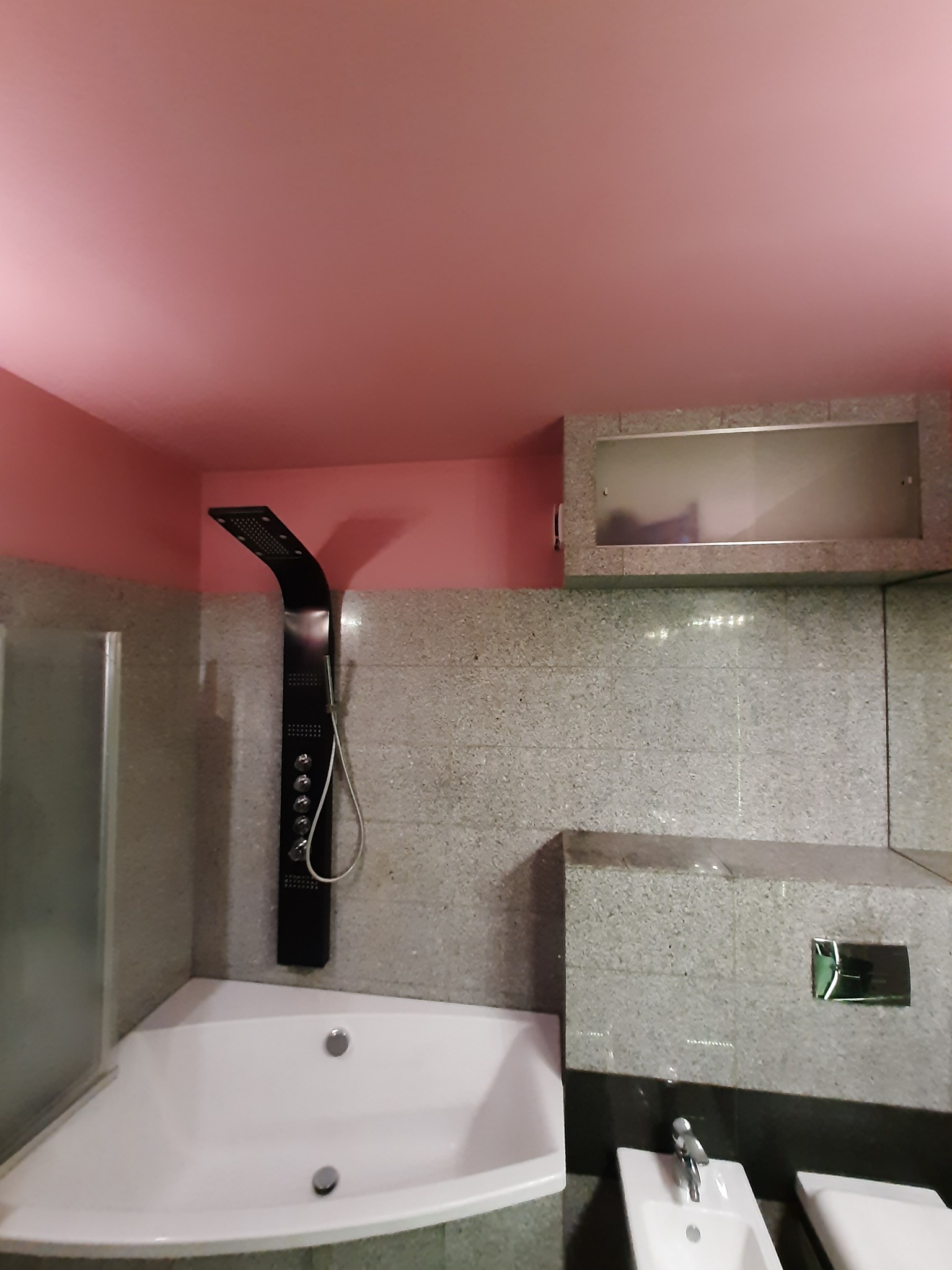 Nowoczesna łazienka z narożną wanną, czarną kolumną prysznicową i bidetem, wykończona płytkami w odcieniach szarości, z różowym sufitem.