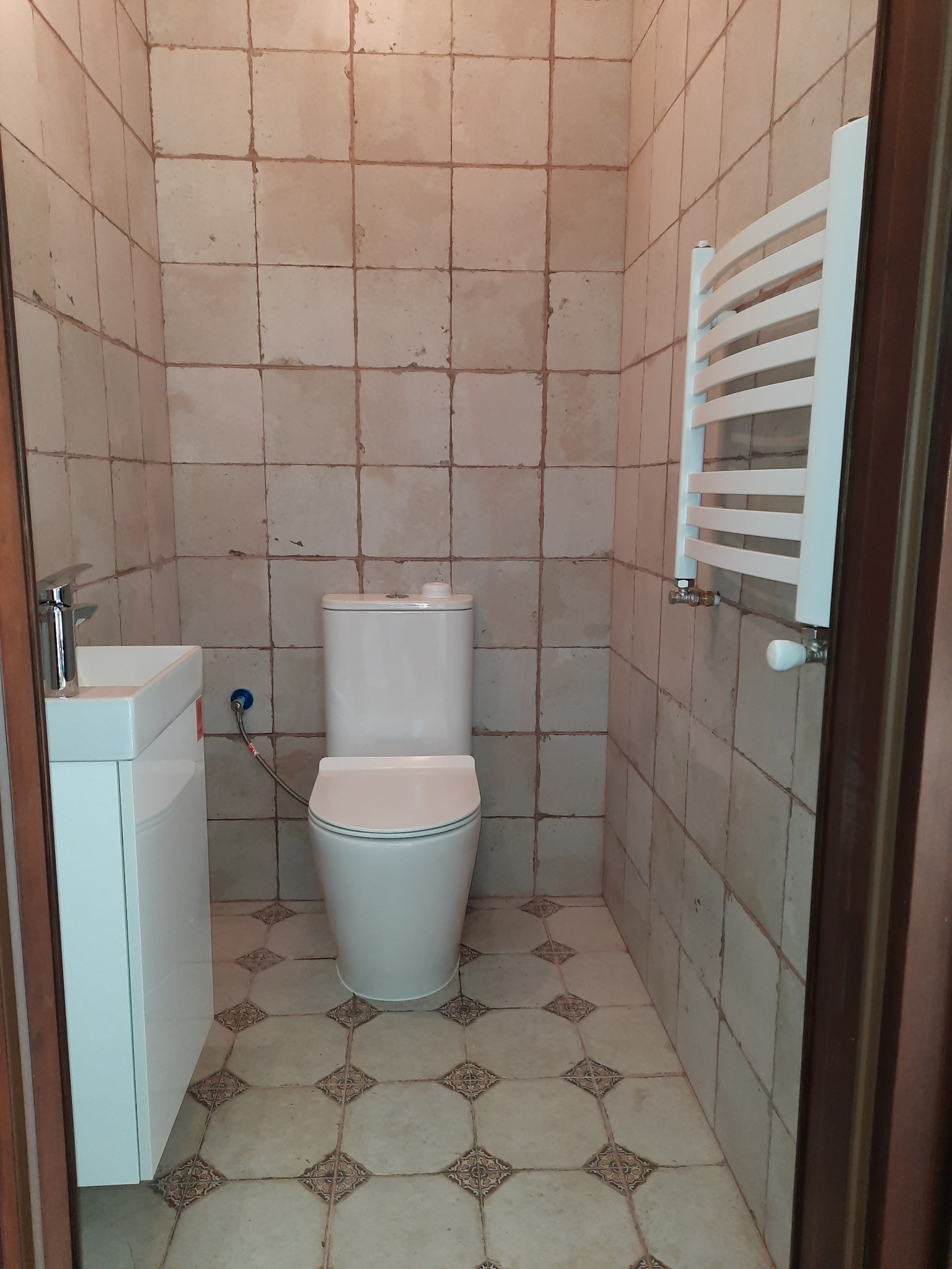 Ukończona łazienka z białą toaletą, umywalką z szafką i grzejnikiem drabinkowym na tle kwadratowych, beżowych płytek ściennych i podłogowych z wzorem.