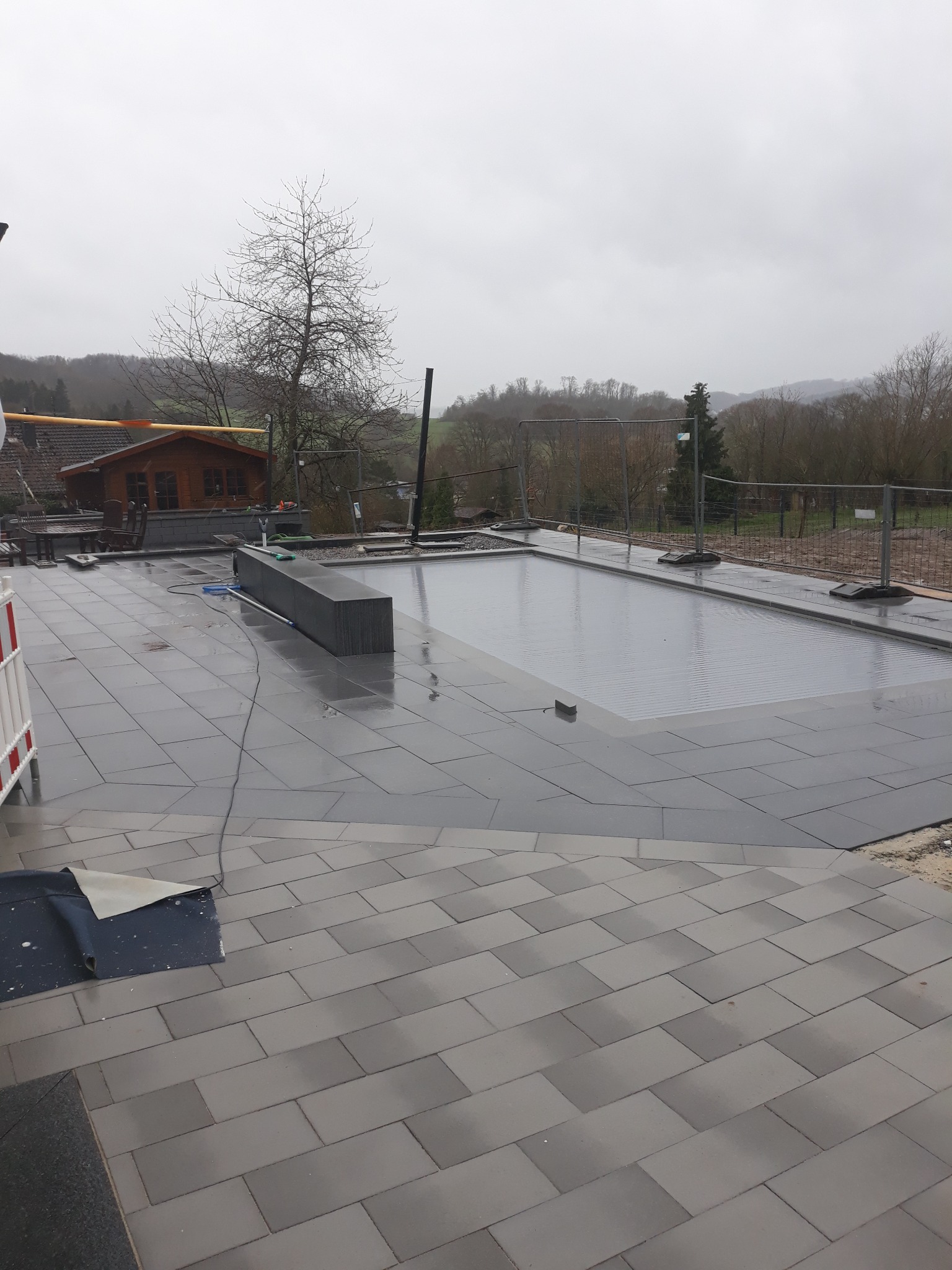 Układanie szarej kostki brukowej wokół basenu z automatyczną roletą, widoczny fragment drewnianego domku i drzewa w tle.