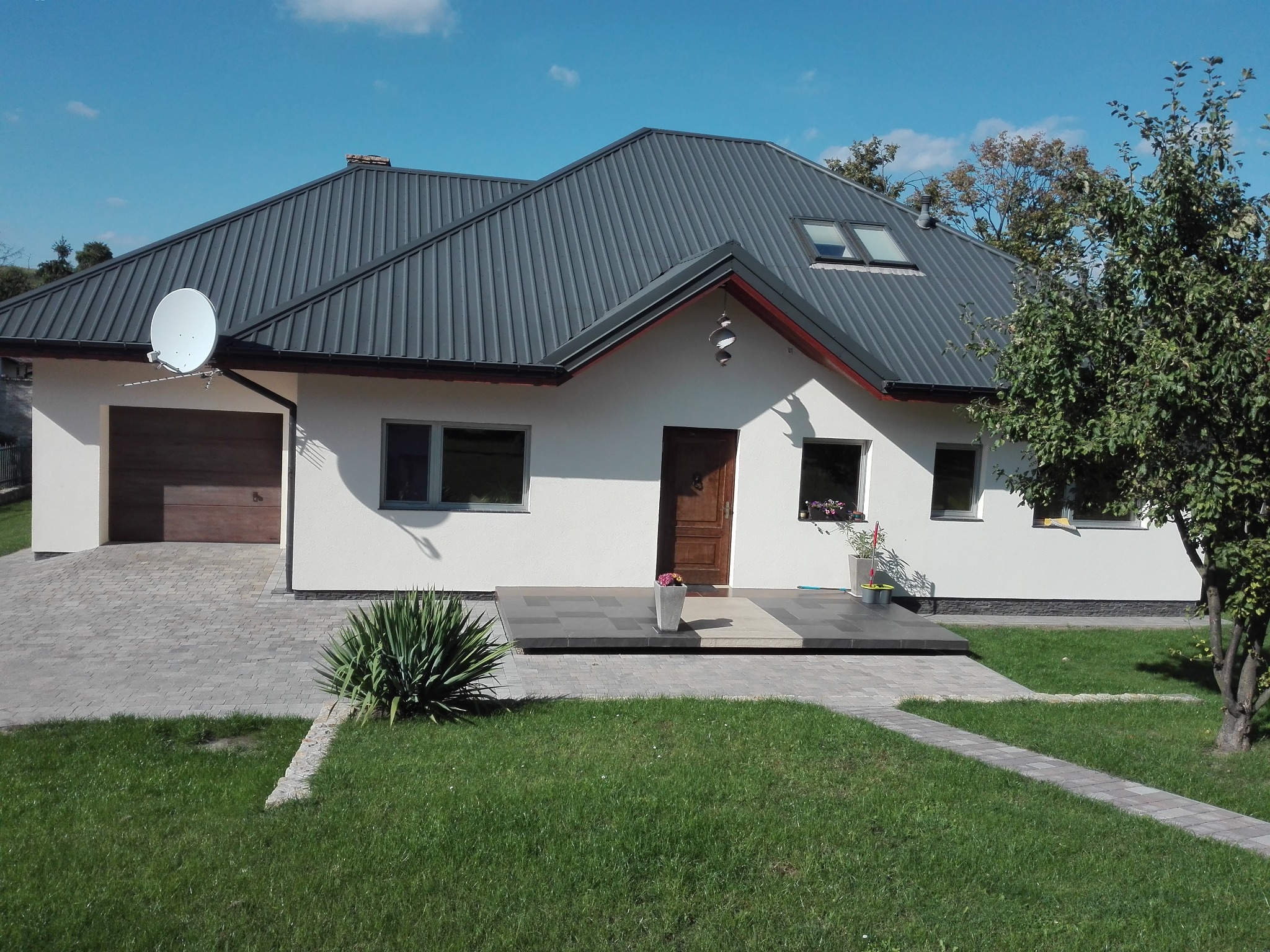 Dom jednorodzinny z szarą blachodachówką, widoczna brama garażowa, drzwi wejściowe z wybrukowanym podejściem i zadbany trawnik.