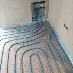 Niebieskie rury ogrzewania podłogowego ułożone na folii z siatką, widoczne kolektory w ścianie w surowym stanie.