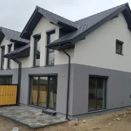 Dwukondygnacyjny dom bliźniak z szarą dachówką, jasną elewacją górnej części i ciemnoszarą dolnej, z szarymi ramami okiennymi i drewnianym ażurowym ogrodzeniem tarasu.