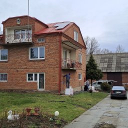 KOZAK Usługi Remontowo Wykończeniowe oraz Budowlane - Dwupiętrowy dom z czerwonej cegły z balkonami; robotnicy ocieplają ścianę zewnętrzną styropianem, jeden stoi na styropianie i nakłada klej, drugi przygotowuje klej na ziemi.