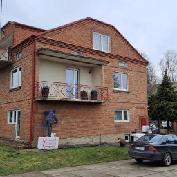 KOZAK Usługi Remontowo Wykończeniowe oraz Budowlane - Mężczyzna w kombinezonie, stojąc na styropianowych płytach, wykonuje prace na elewacji dwukondygnacyjnego, ceglanego domu z balkonami, widoczny samochód osobowy i bus.