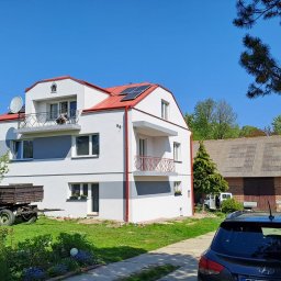 KOZAK Usługi Remontowo Wykończeniowe oraz Budowlane - Nowoczesny, dwupiętrowy dom z balkonami, czerwonym dachem i panelami słonecznymi, widoczny z poziomu podjazdu, z przyczepą rolniczą i budynkiem gospodarczym w tle.