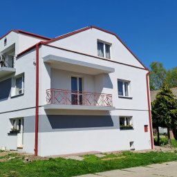 KOZAK Usługi Remontowo Wykończeniowe oraz Budowlane - Dwupiętrowy dom z balkonami, pomalowany na biało z szarymi pasami i czerwonymi rynnami, na jednym z balkonów stoi mężczyzna, na podwórku zaparkowany samochód dostawczy.