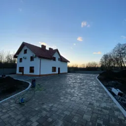 Nowy podjazd z szarej kostki brukowej ułożonej we wzór jodełki przed dwupiętrowym domem z czerwoną dachówką, widoczna myjka ciśnieniowa z zielonym wężem, świeżo nasypana ziemia po bokach.