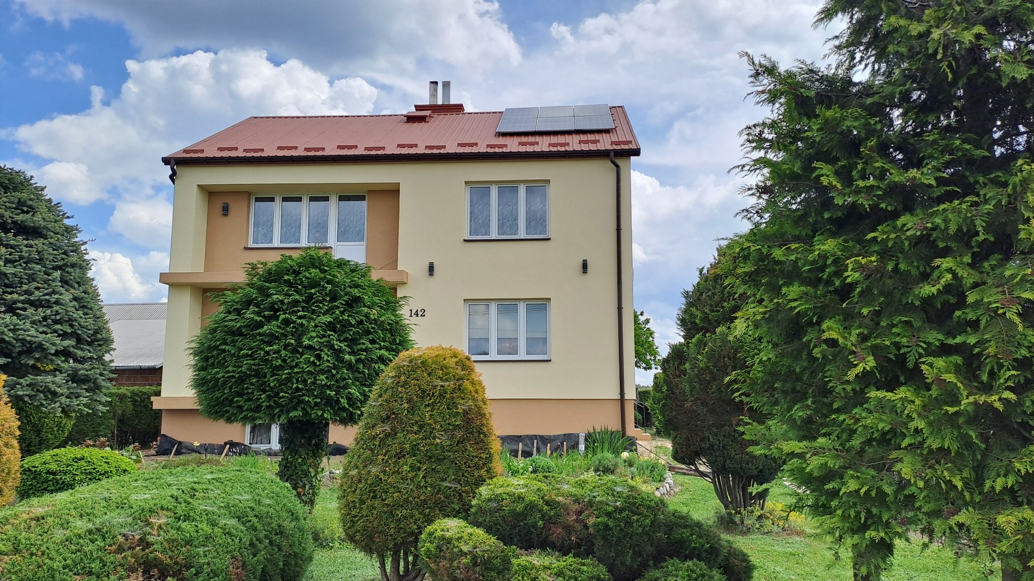 Dwukondygnacyjny dom z beżową elewacją, czerwoną dachówką i panelami słonecznymi, otoczony ozdobnymi krzewami i drzewami w słoneczny dzień.