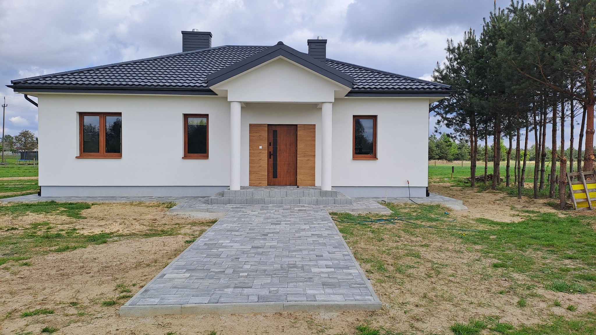 Nowoczesny dom jednorodzinny z szarą kostką brukową prowadzącą do wejścia, widoczne drewniane okna i drzwi, oraz jasna elewacja i ciemny dach.