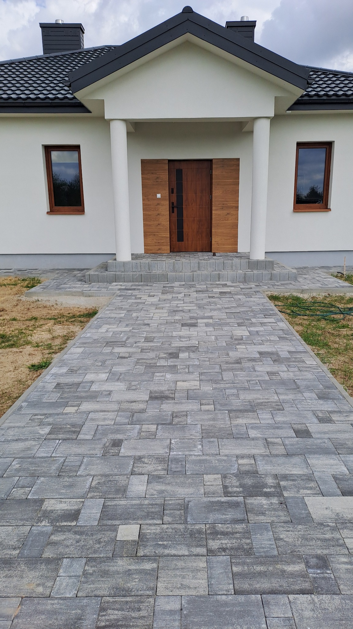 Nowoczesny podjazd z szarej kostki brukowej prowadzący do wejścia domu z drewnianymi drzwiami i białymi kolumnami.