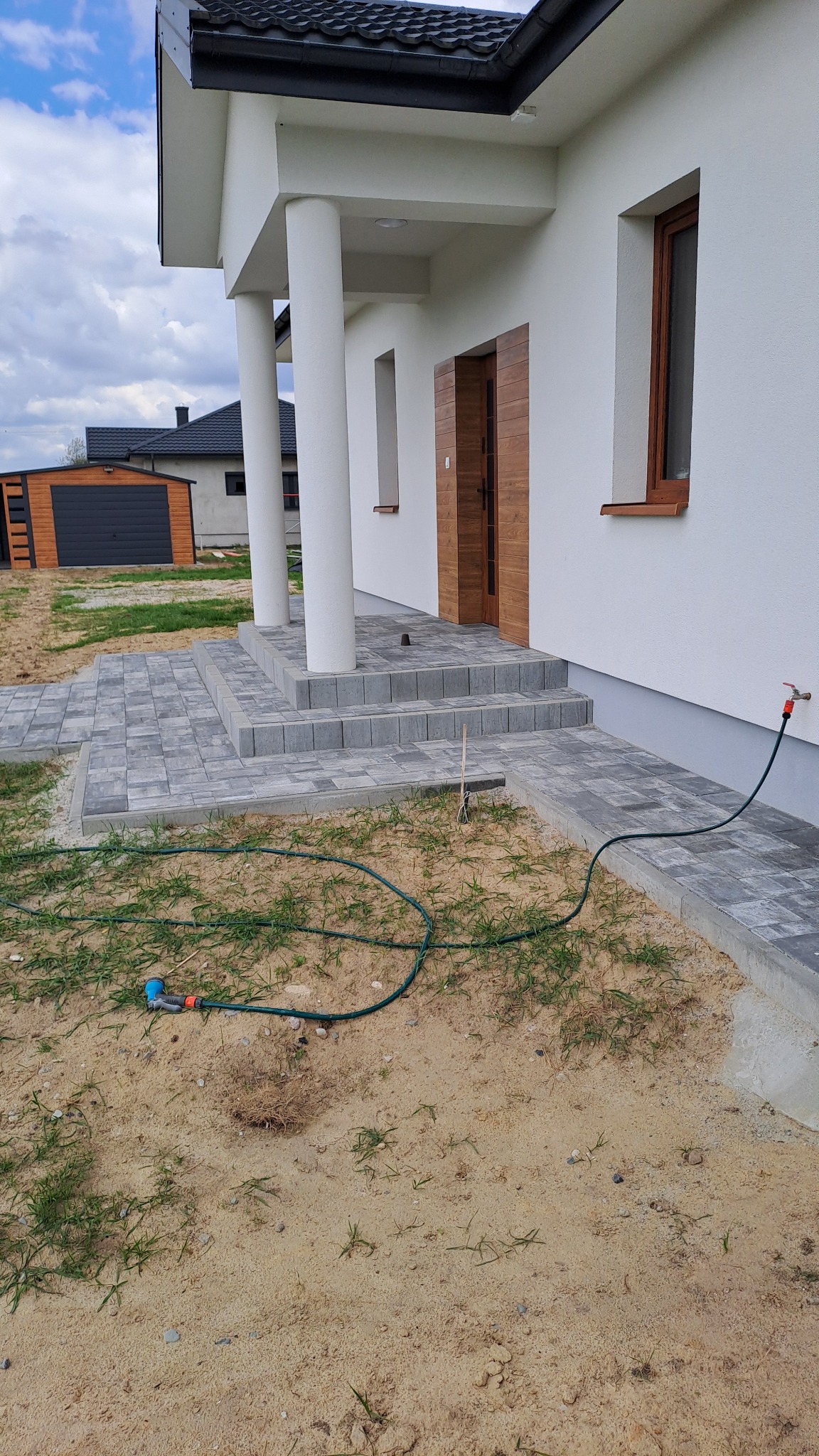 Ułożona kostka brukowa prowadząca do wejścia domu z kolumnami, widoczny wąż ogrodowy i piaszczyste podłoże z kępkami trawy.