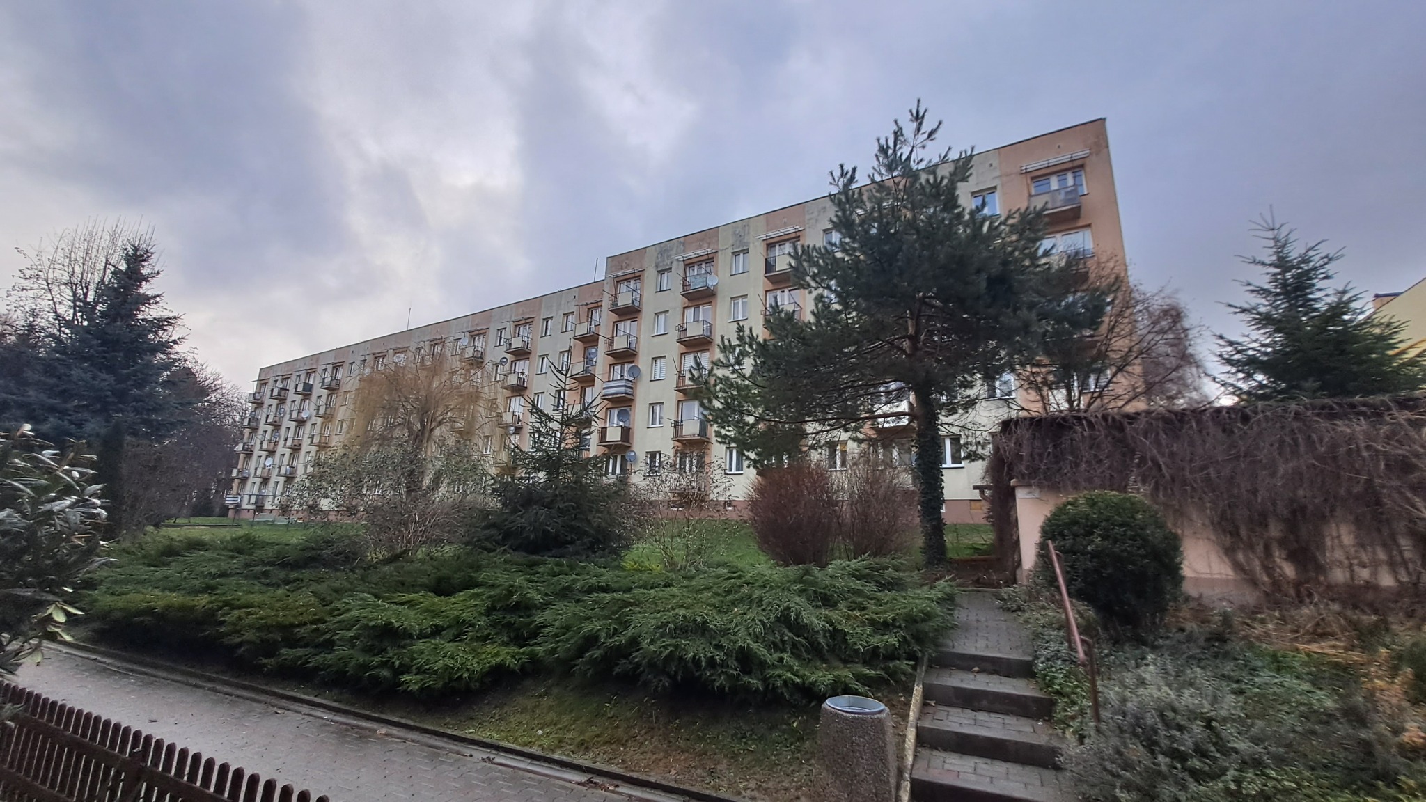 Wielopiętrowy budynek mieszkalny z balkonami, otoczony zielenią i drzewami w pochmurny dzień.