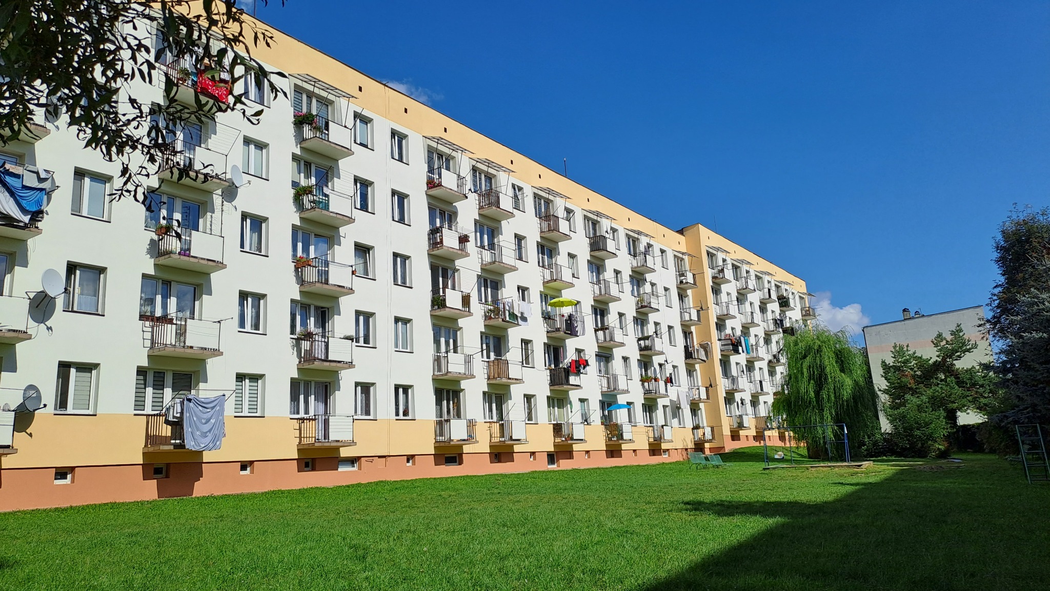 Wielopiętrowy budynek mieszkalny z balkonami, świeżo pomalowaną elewacją w kolorach bieli, żółci i brązu, widoczny z perspektywy trawnika w słoneczny dzień.
