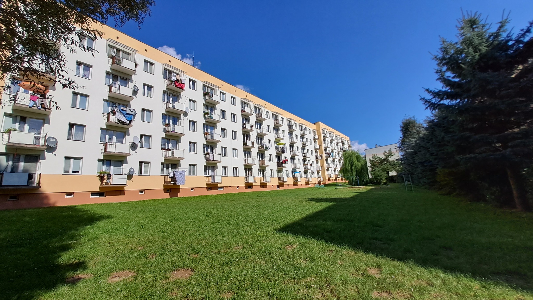 Wielopiętrowy budynek mieszkalny z balkonami, widok z trawnika w słoneczny dzień. Na balkonach suszące się pranie i kwiaty w doniczkach.