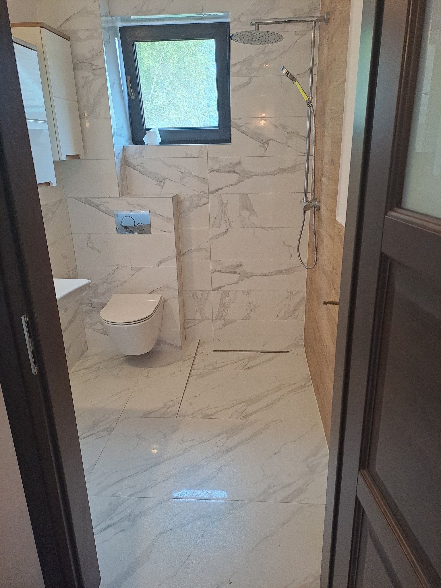 Nowoczesna łazienka z białymi marmurkowymi płytkami, wiszącą toaletą i kabiną prysznicową typu walk-in z odpływem liniowym, widziana zza uchylonych, ciemnych drzwi.