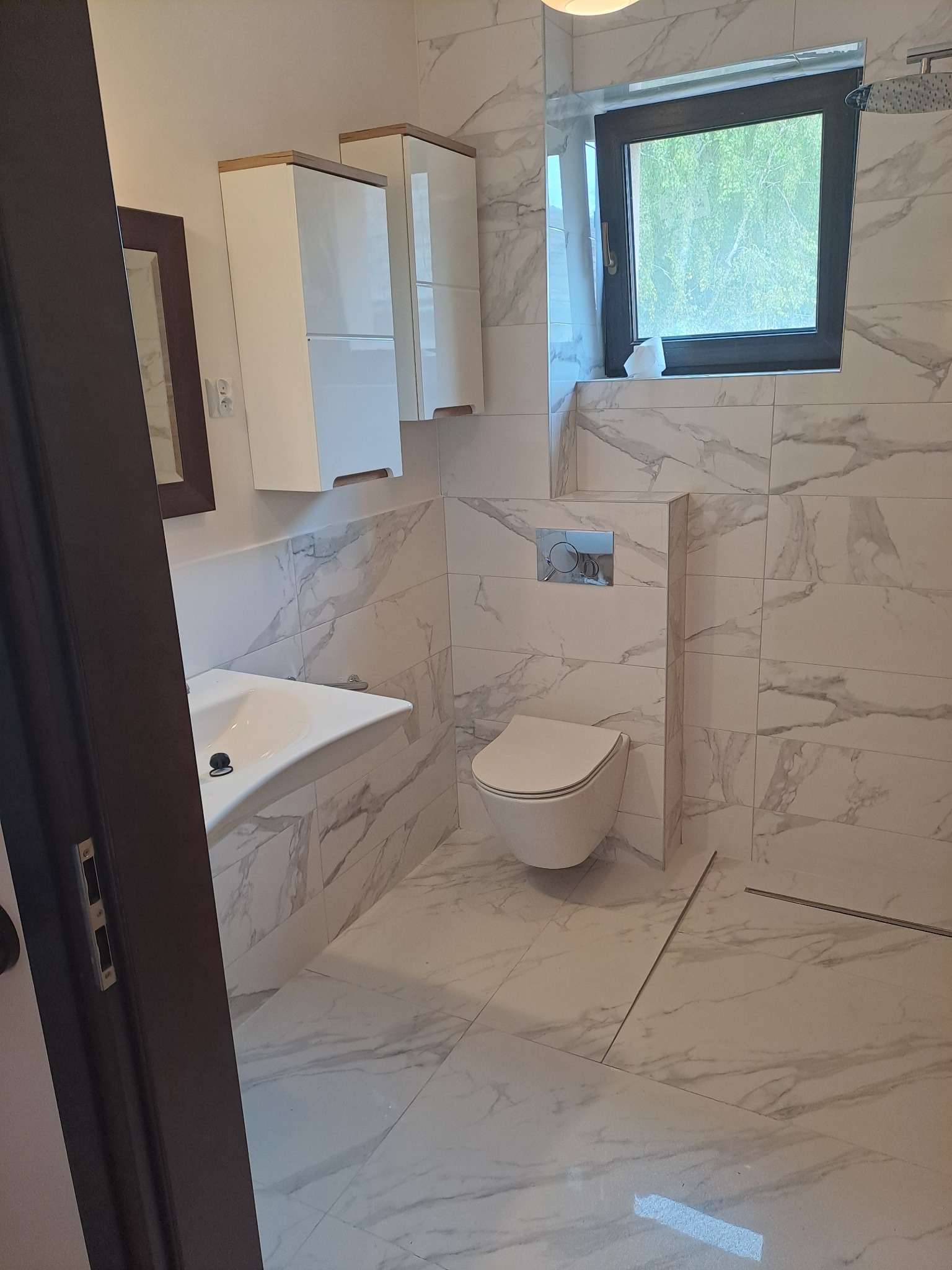 Nowoczesna łazienka z marmurowymi płytkami, wiszącą toaletą i szafkami w kolorze białym z drewnianymi akcentami, widok na okno z zielenią na zewnątrz.