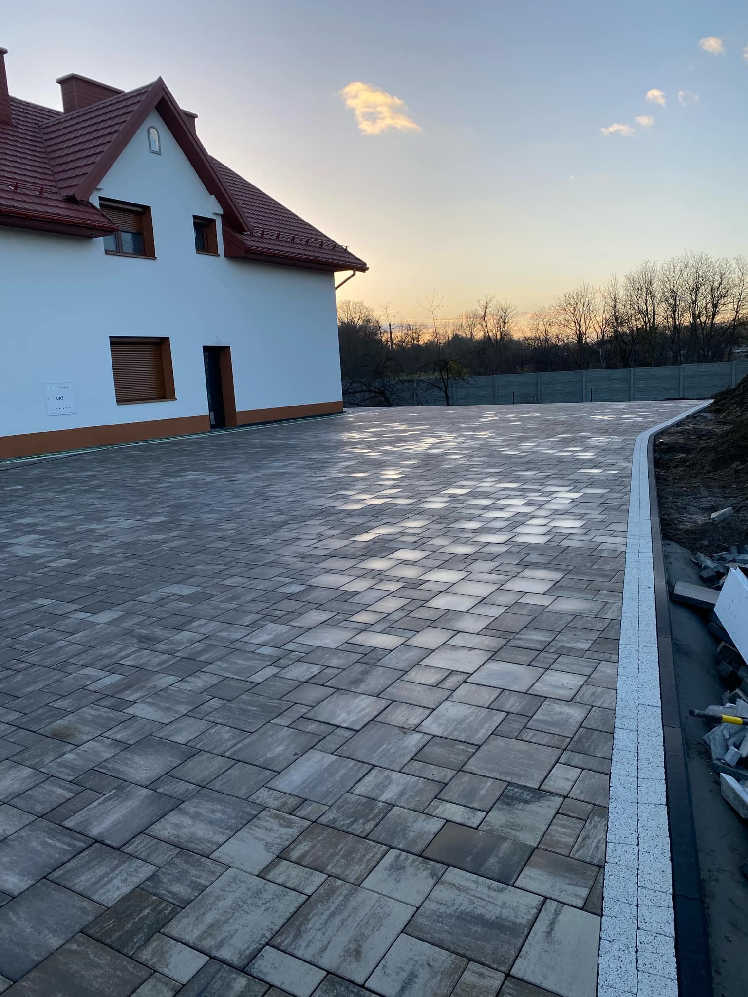 Szeroki podjazd z szarej kostki brukowej ułożonej we wzór, obrzeża wykończone białą kostką, dom z czerwonym dachem w tle, widok na zachodzące słońce.