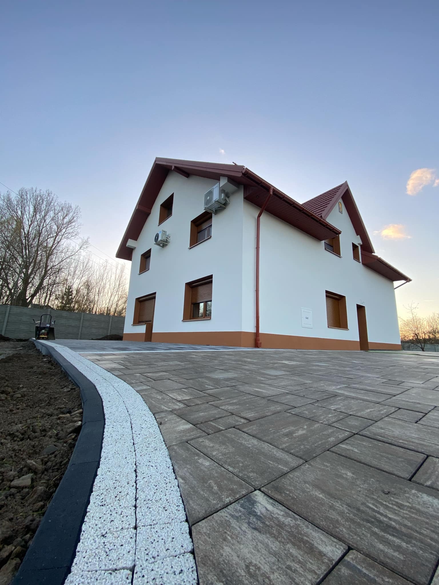 Ułożona szara kostka brukowa prowadząca do nowoczesnego, białego domu z brązowymi akcentami i klimatyzacją, widok z niskiej perspektywy, obrzeże z białego kruszywa.