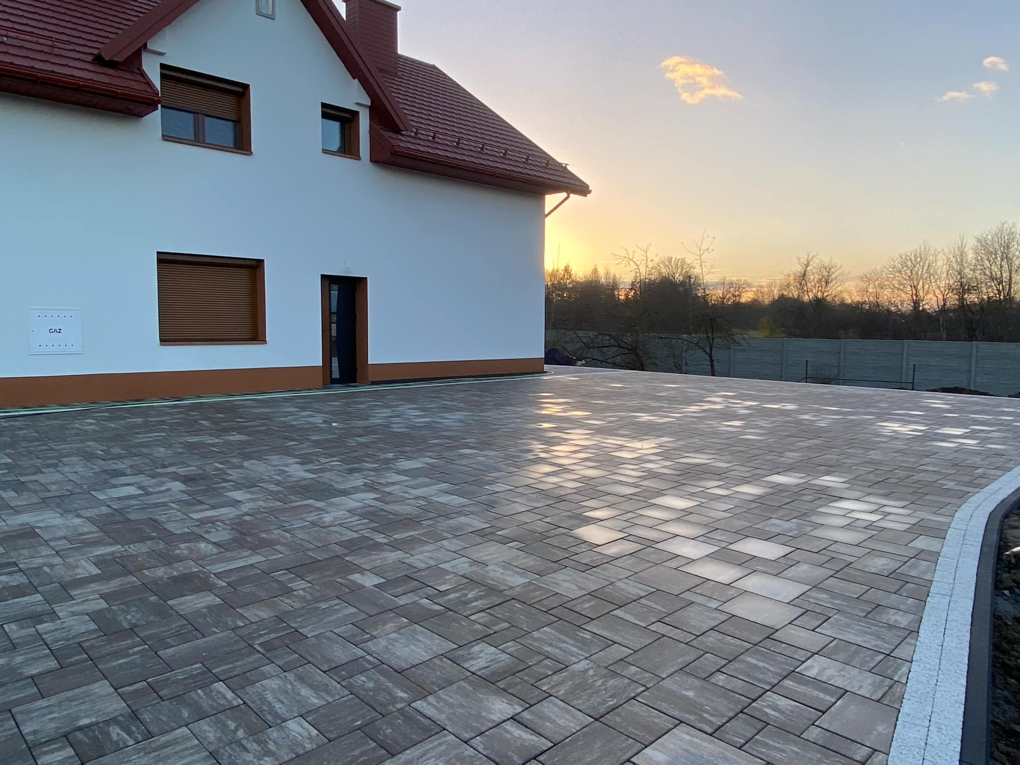 Nowoczesna posesja z nowo ułożoną, szarą kostką brukową w geometryczny wzór, obrzeża wykończone jasnym kamieniem, dom z białą elewacją i czerwoną dachówką w tle, zachodzące słońce.