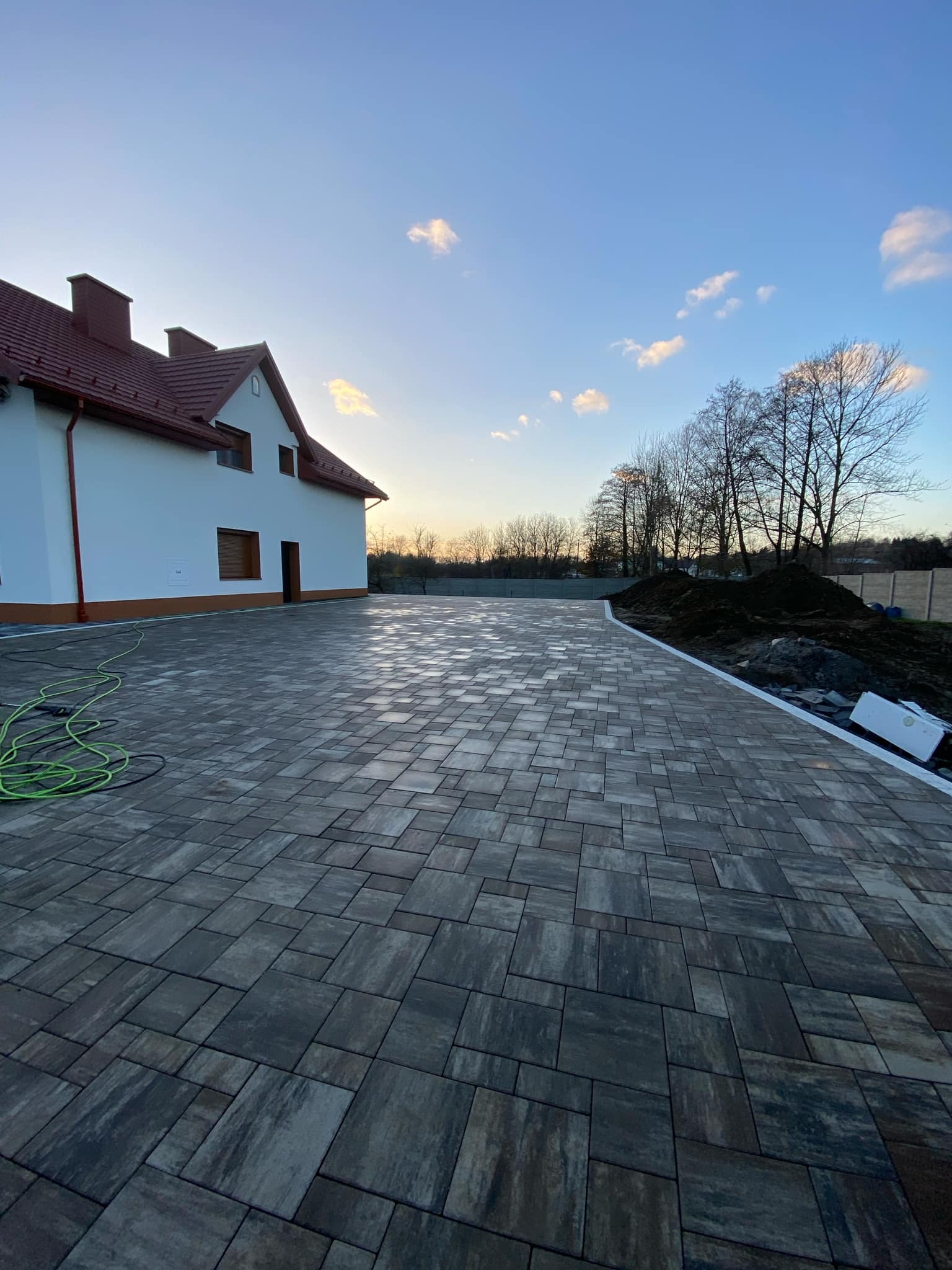 Ułożona kostka brukowa w odcieniach szarości na posesji przed domem jednorodzinnym, widoczny fragment domu z czerwoną dachówką i zielony wąż ogrodowy.