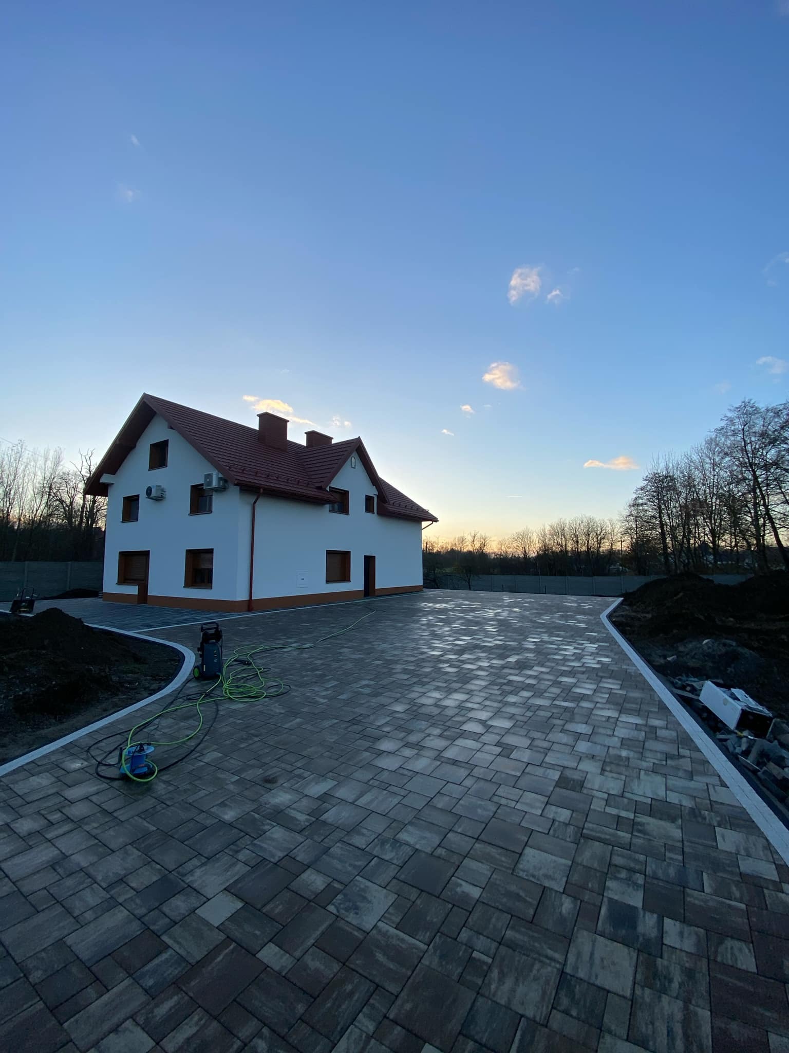 Nowy podjazd z szarej kostki brukowej ułożonej we wzór jodełki przed dwupiętrowym domem z czerwoną dachówką, widoczna myjka ciśnieniowa z zielonym wężem, świeżo nasypana ziemia po bokach.