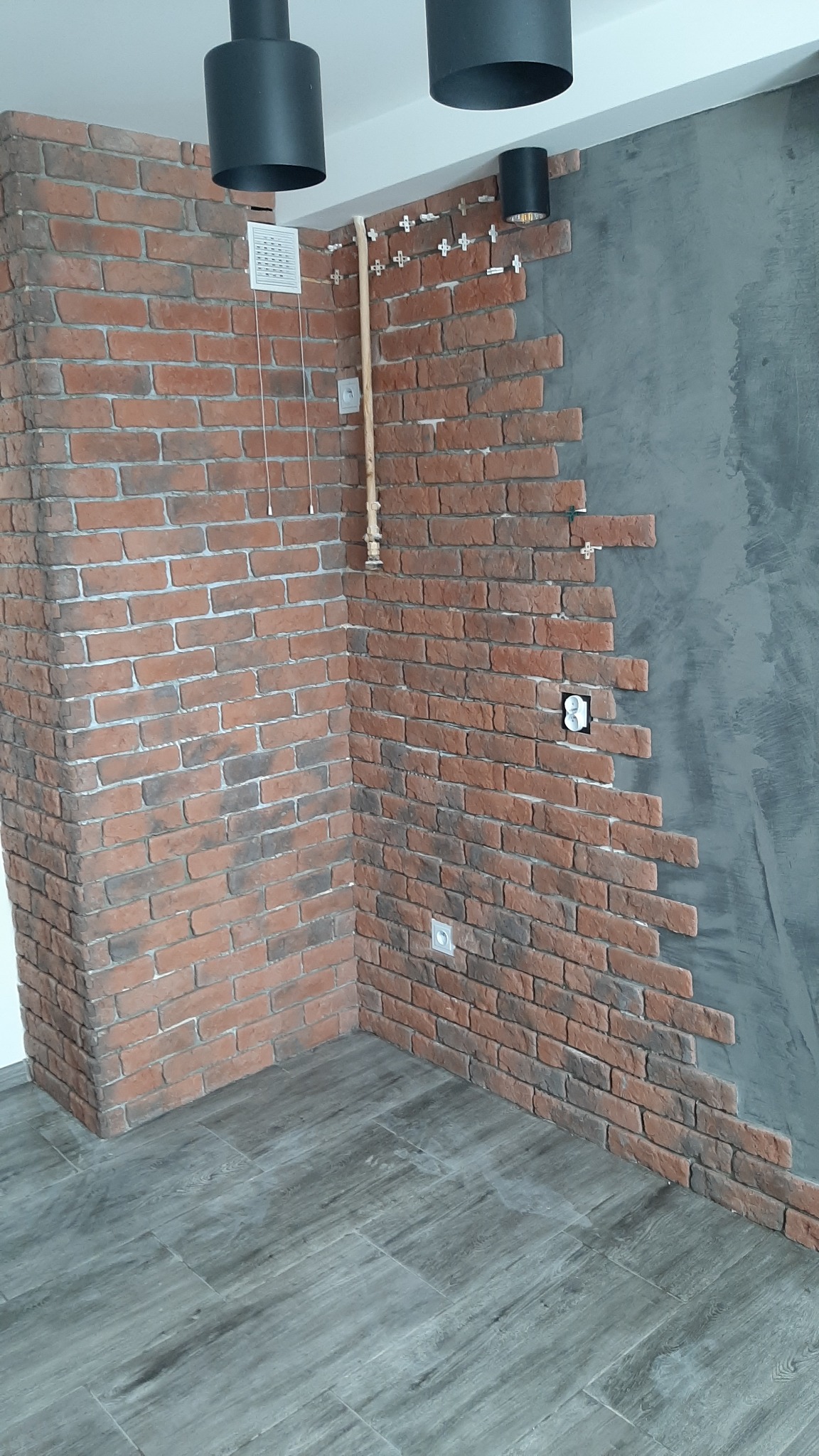 Fragment ściany z częściowo ułożoną czerwoną cegłą dekoracyjną, widoczne krzyżyki dystansowe, instalacja elektryczna i fragment szarej ściany, podwieszane lampy sufitowe.