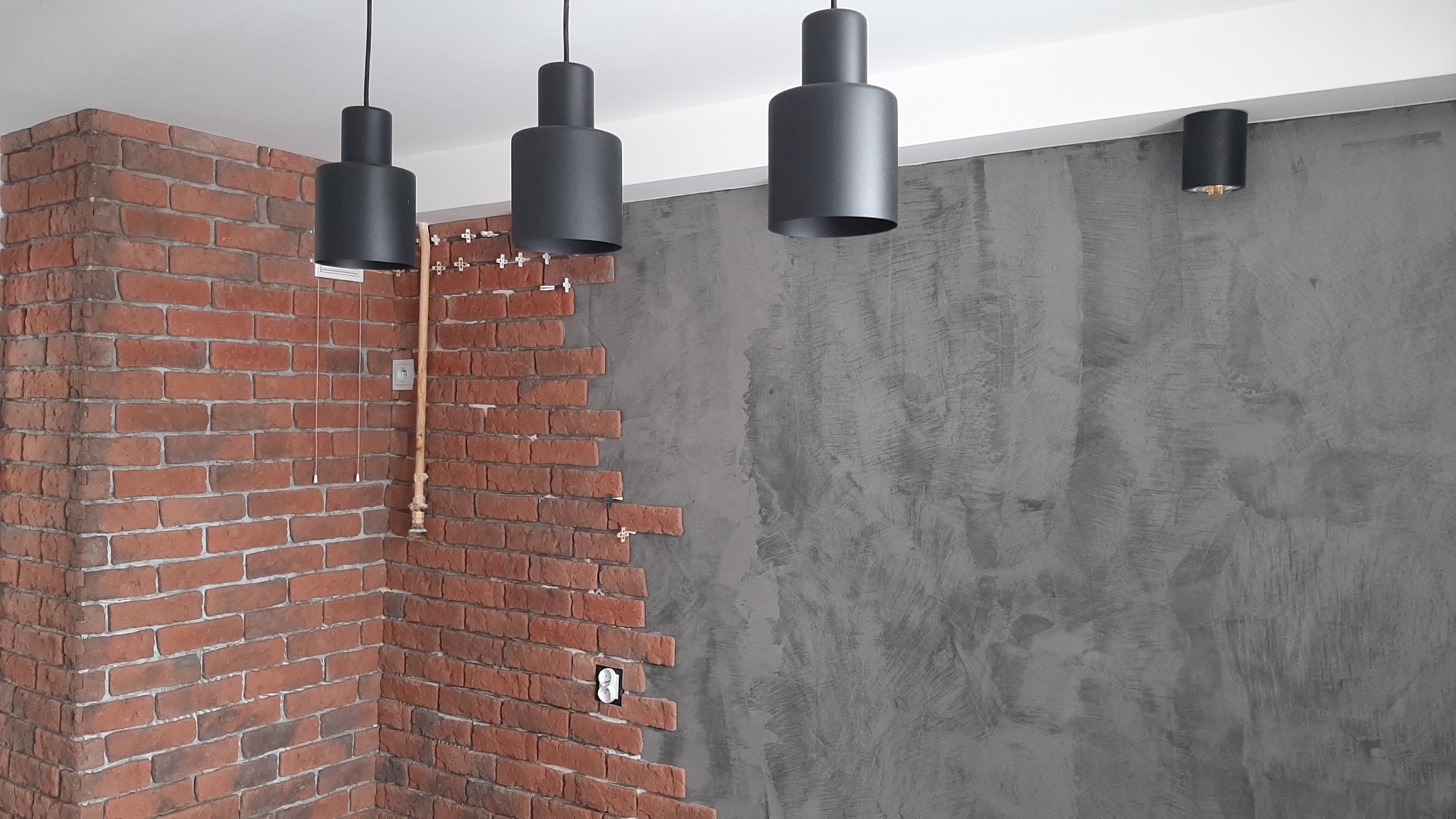 Wnętrze w trakcie wykańczania: częściowo ułożona okładzina z cegły na ścianie, surowa szara ściana obok, wiszące czarne lampy tuby i mały kinkiet na szarej ścianie.