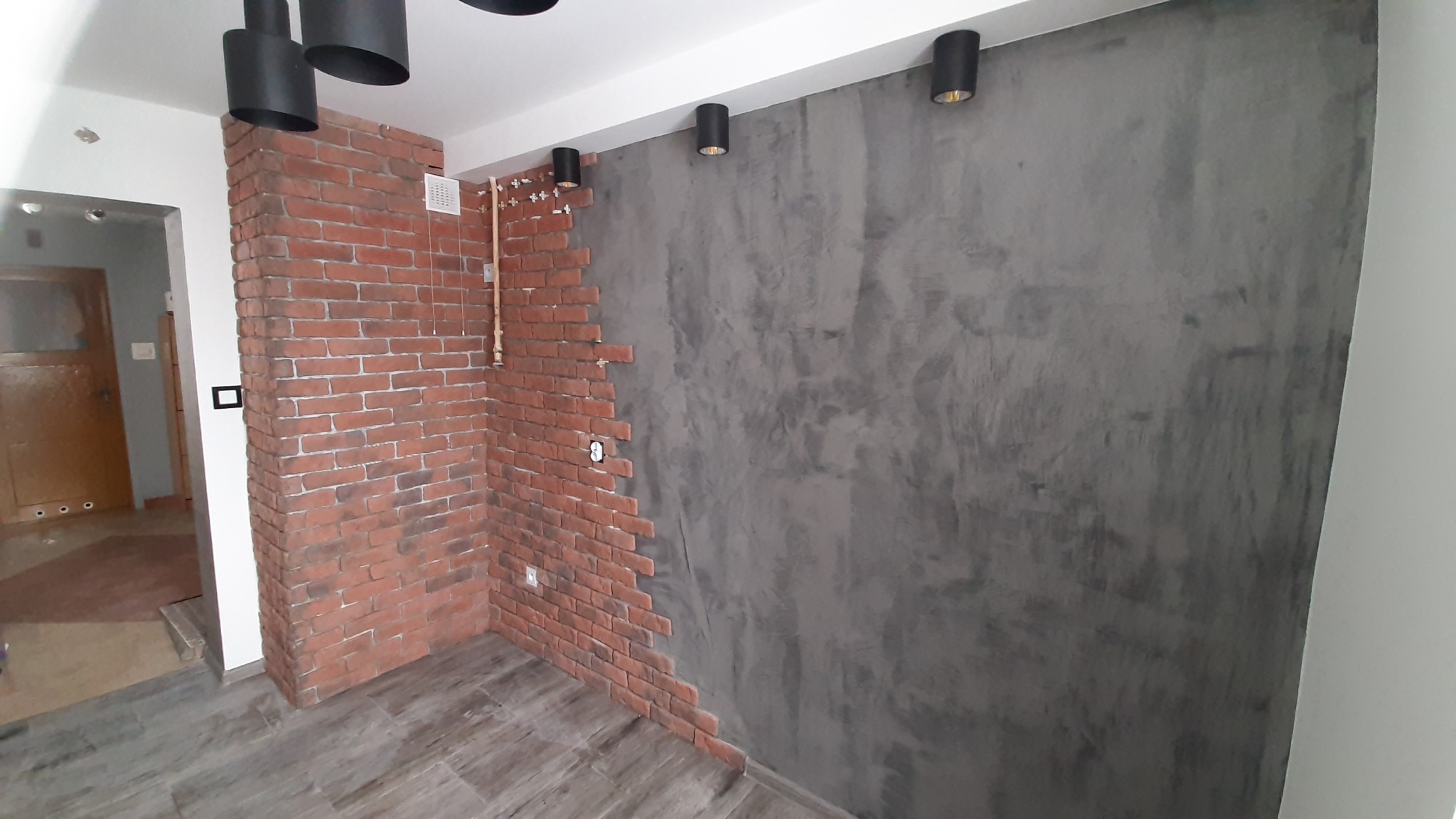 Wnętrze w trakcie wykończenia: fragment ściany z dekoracyjną cegłą, sąsiadujący z szarą, fakturowaną ścianą. Widoczne czarne lampy sufitowe i instalacja elektryczna.