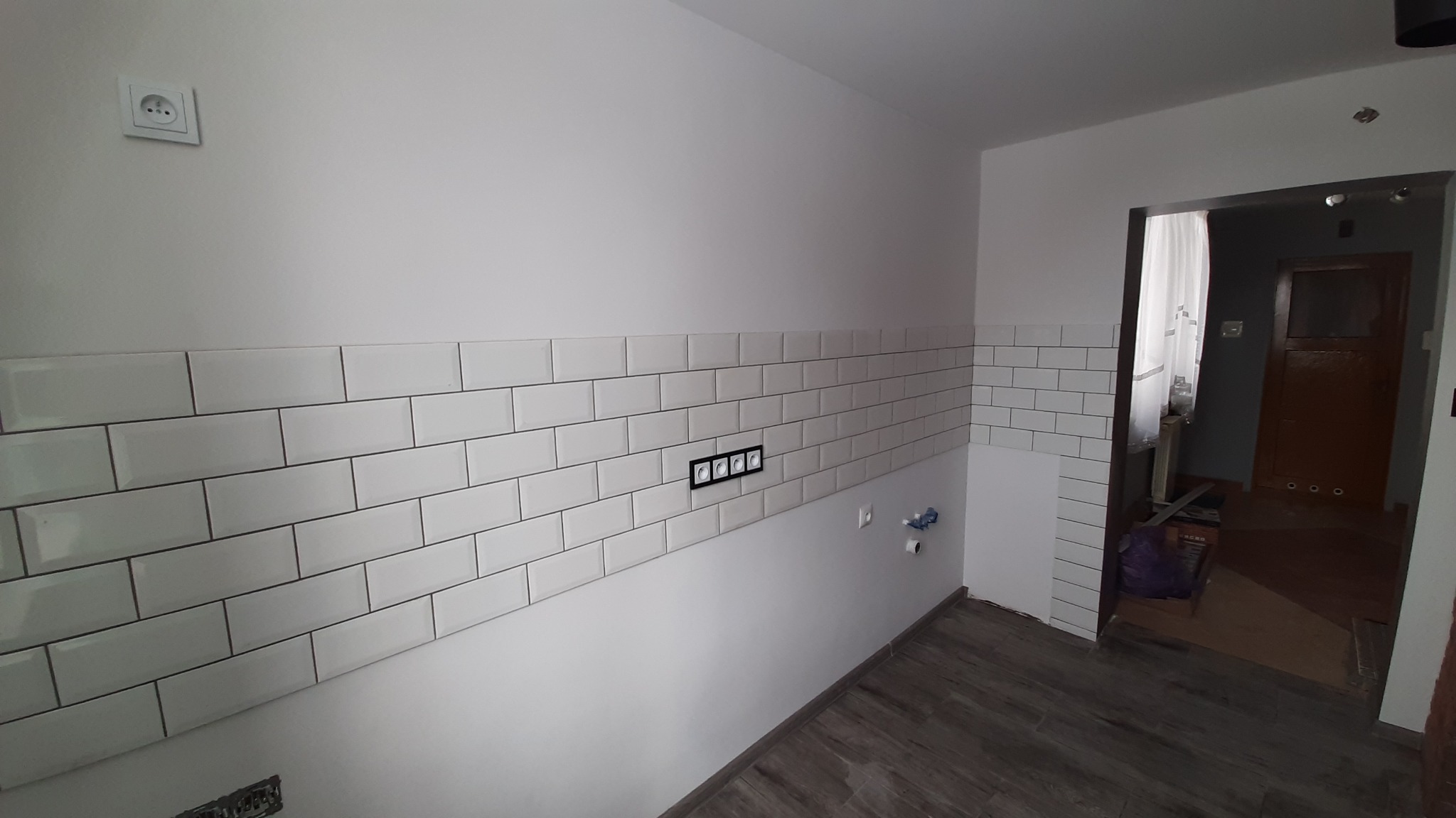 Ściana w trakcie wykańczania z białymi, prostokątnymi płytkami, widoczne gniazdka elektryczne w czarnej ramce, szare panele podłogowe i otwarte przejście do innego pomieszczenia.