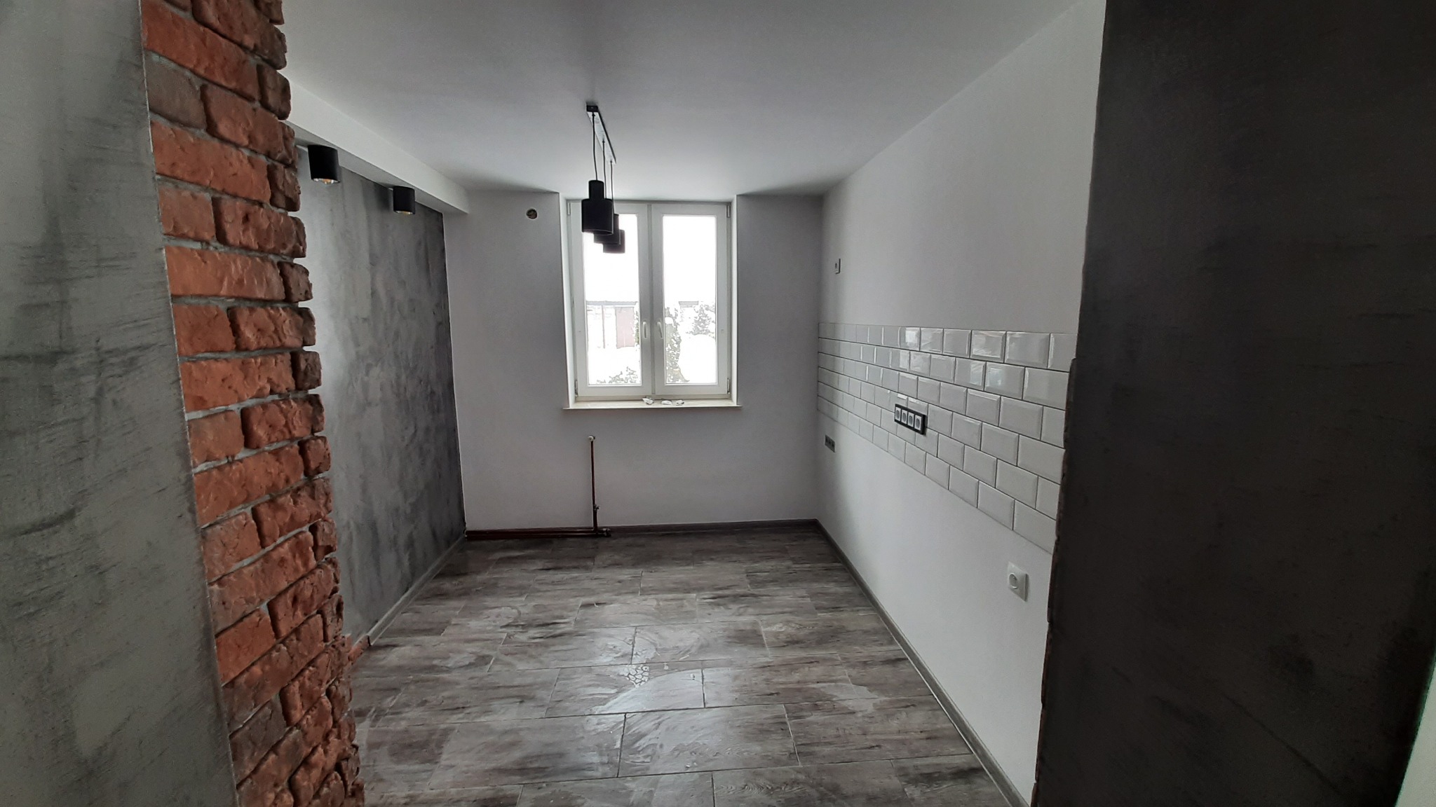 Surowe wnętrze po remoncie: ceglana ściana, szare tynki dekoracyjne, białe płytki metra na ścianie, drewnopodobna podłoga, czarne lampy wiszące