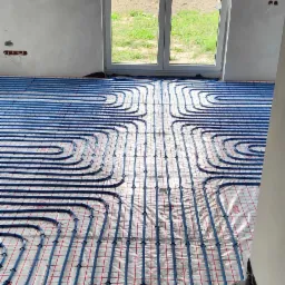 Niedokończona instalacja ogrzewania podłogowego z niebieskimi rurkami na folii z siatką w nowym budynku, widok na okno z zielonym trawnikiem na zewnątrz.