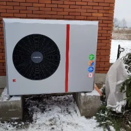 Szara pompa ciepła Kolton z czerwoną linią, umieszczona na betonowych blokach przed ceglaną ścianą budynku w zimowej scenerii, widoczne naklejki z klasą energetyczną i czynnikiem chłodniczym R290.