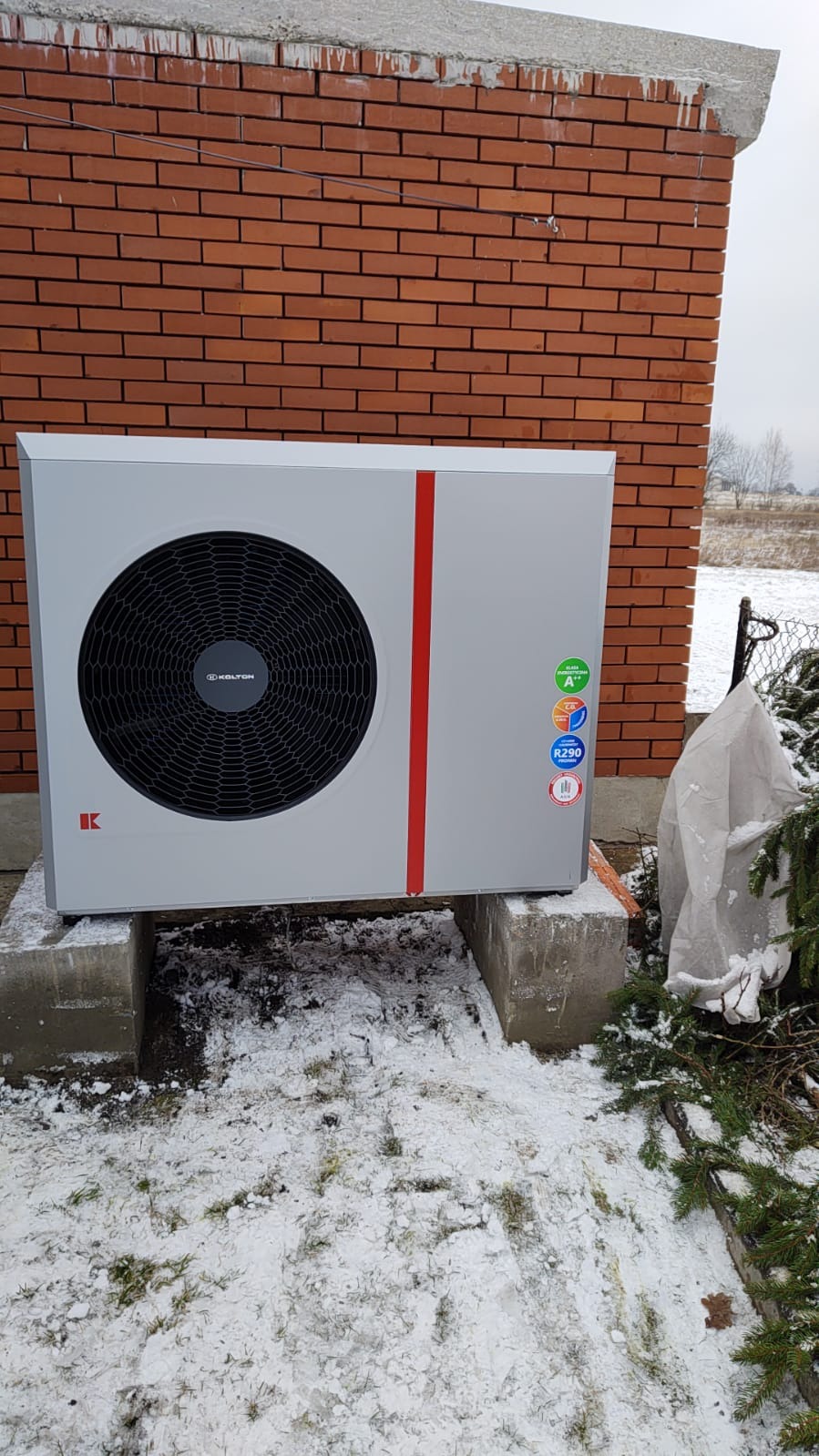 Szara pompa ciepła Kolton z czerwoną linią, umieszczona na betonowych blokach przed ceglaną ścianą budynku w zimowej scenerii, widoczne naklejki z klasą energetyczną i czynnikiem chłodniczym R290.