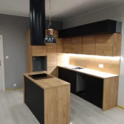 Nowoczesna kuchnia z wyspą, fronty szafek w kolorze drewna z czarnymi akcentami, oświetlenie LED pod szafkami wiszącymi, czarny okap cylindryczny i lampa wisząca z elementami miedzianymi.