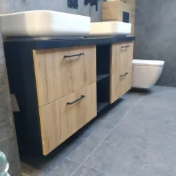 Nowoczesna łazienka z dwoma białymi umywalkami nablatowymi na szafce z drewnopodobnymi frontami i czarnym blatem, szare płytki na ścianach i podłodze, wisząca toaleta w tle.