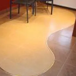 Beżowa podłoga z płytek o nieregularnym kształcie, oddzielona od brązowych płytek dekoracyjną listwą, widoczne krzesła i fragment ściany w tle.