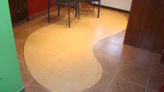 Beżowa podłoga z płytek o nieregularnym kształcie, oddzielona od brązowych płytek dekoracyjną listwą, widoczne krzesła i fragment ściany w tle.