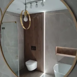 Okrągłe lustro w złotej ramie odbija nowoczesną łazienkę z szarymi płytkami imitującymi marmur, podświetlaną pionową listwą LED, wiszącą toaletą, umywalką i dekoracyjnym oświetleniem punktowym...