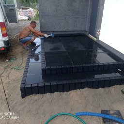 Mój raj - Mężczyzna w trakcie układania czarnych, błyszczących płyt na schodach wejściowych do budynku, używa gumowego młotka, widoczny samochód dostawczy w tle.