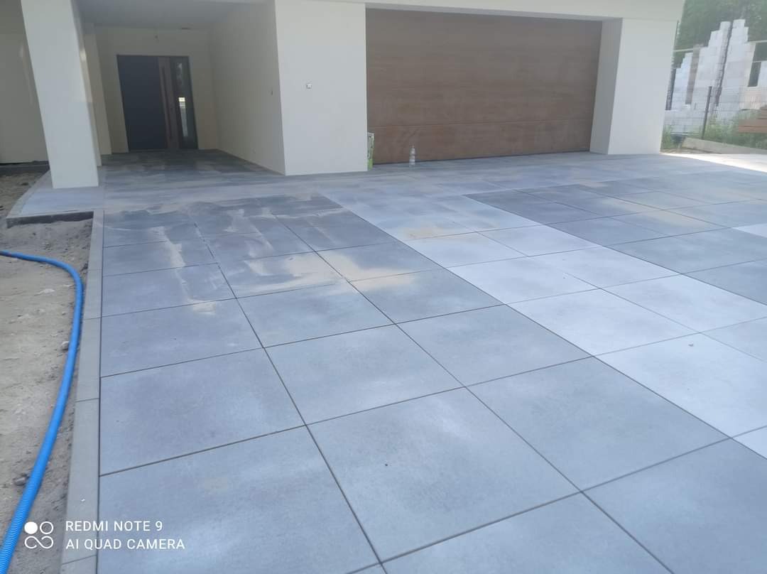 Ułożona szara kostka brukowa na podjeździe do garażu nowoczesnego domu, widoczny fragment niebieskiego węża ogrodowego.