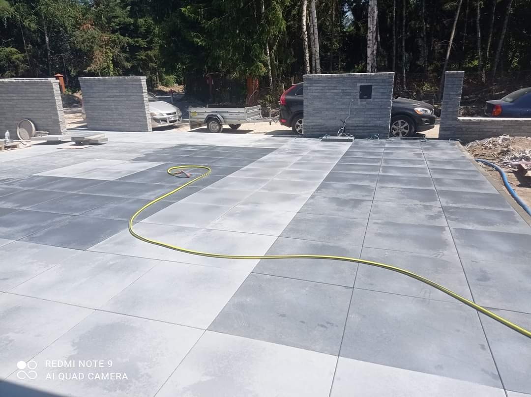 Ułożona nawierzchnia z szarych płyt betonowych w dwóch odcieniach, z widocznymi elementami ogrodzenia z tego samego materiału w tle oraz zaparkowanymi samochodami i przyczepką.