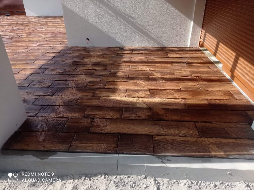 Ułożona nawierzchnia z kostki brukowej imitującej drewno, z widocznymi słojami i fakturą, jasne ściany budynku w tle i częściowo opuszczona brązowa roleta.