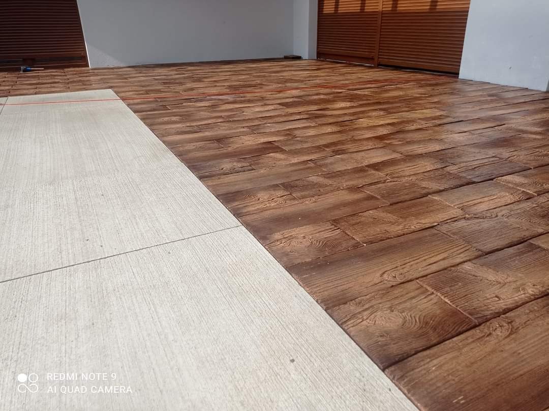 Nowo ułożona, brązowa kostka brukowa imitująca drewno, sąsiadująca z jasną, betonową nawierzchnią, widoczna brama garażowa w tle.