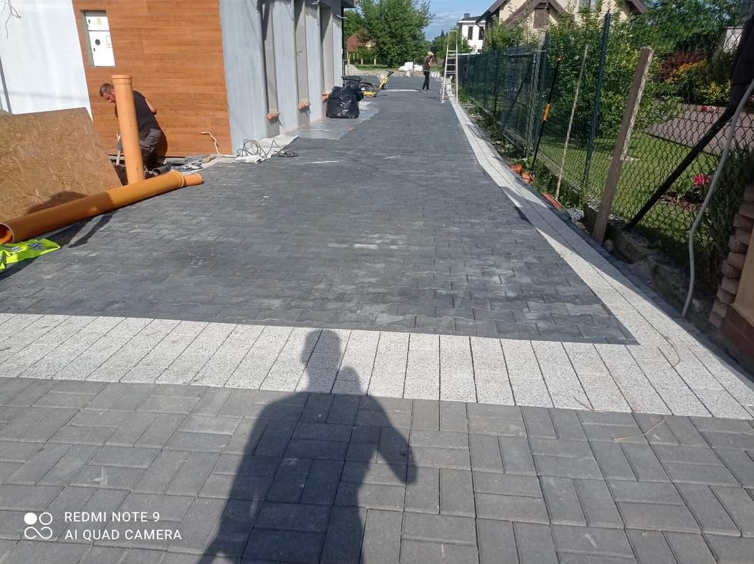 Nowo ułożona, szara kostka brukowa z jasnoszarą obwódką, widoczny pracownik z rurą PCV oraz częściowo ukończona instalacja drenażowa przy budynku.