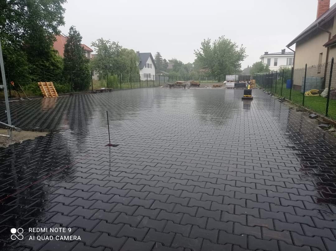 Świeżo ułożona, mokra od deszczu, grafitowa kostka brukowa na posesji, widoczny ubijak wibracyjny, palety z kostką w tle, pochmurny dzień.