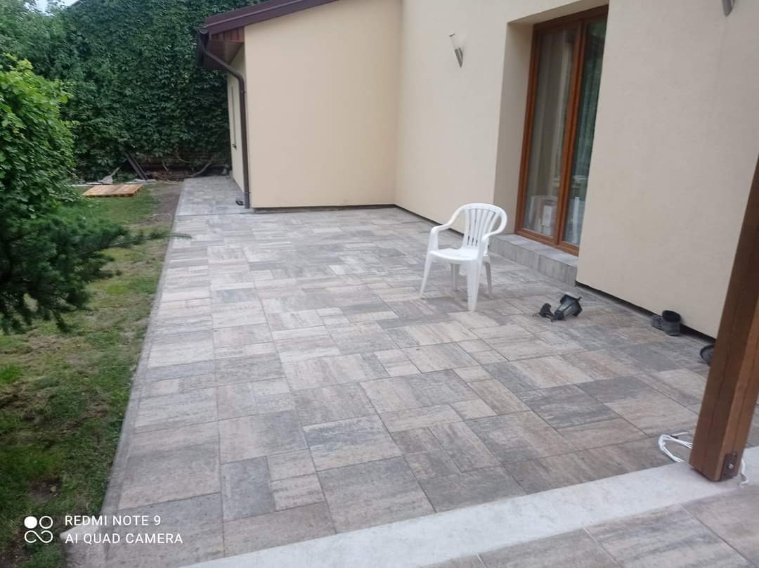 Ułożona szara kostka brukowa na tarasie przydomowym, białe krzesło plastikowe, para butów roboczych i kalosze przy ścianie budynku.