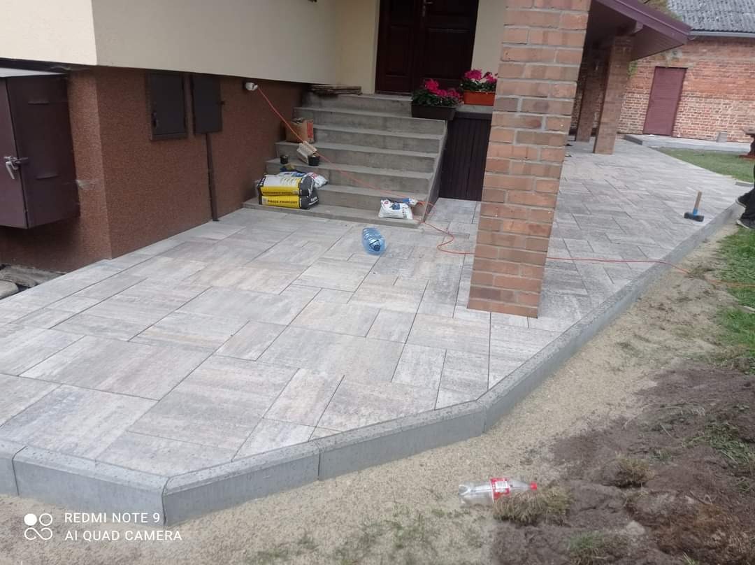 Nowo ułożona, szara kostka brukowa przed domem z ceglaną kolumną i schodami. Widoczne materiały budowlane i butelki.