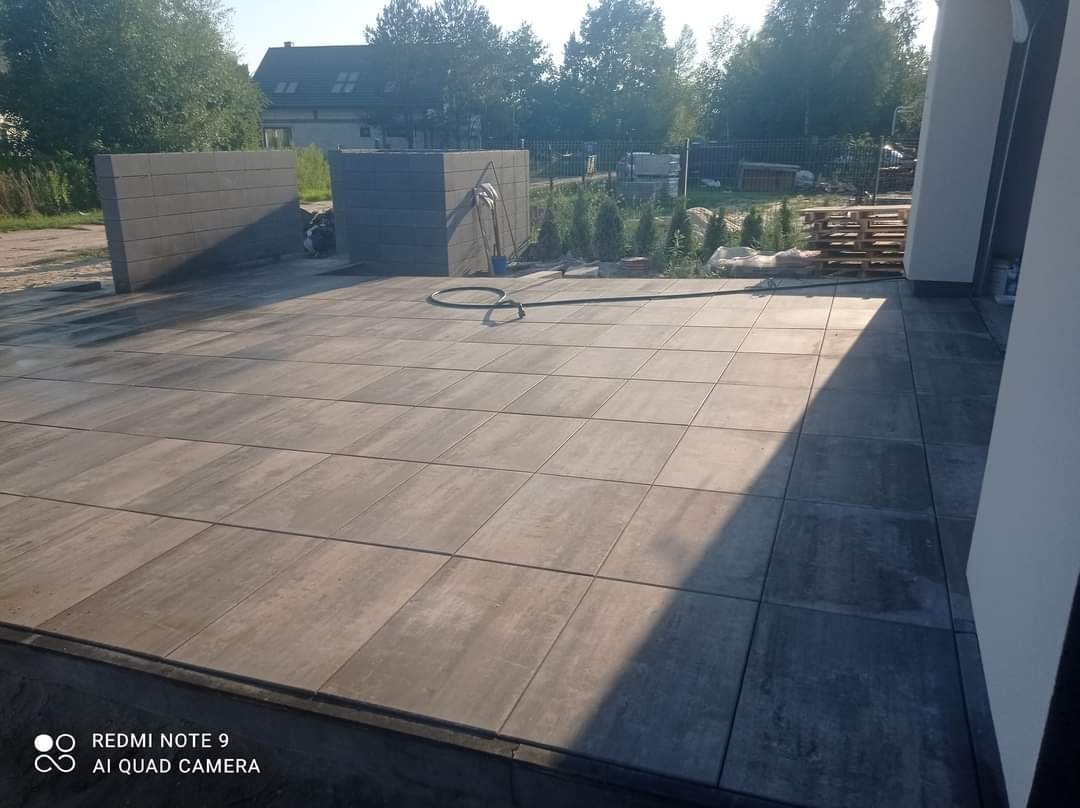 Nowa, szara nawierzchnia z prostokątnych płyt betonowych na posesji, widoczne dwa murki z bloczków betonowych, węże do wody i palety, w tle dom jednorodzinny i zieleń.