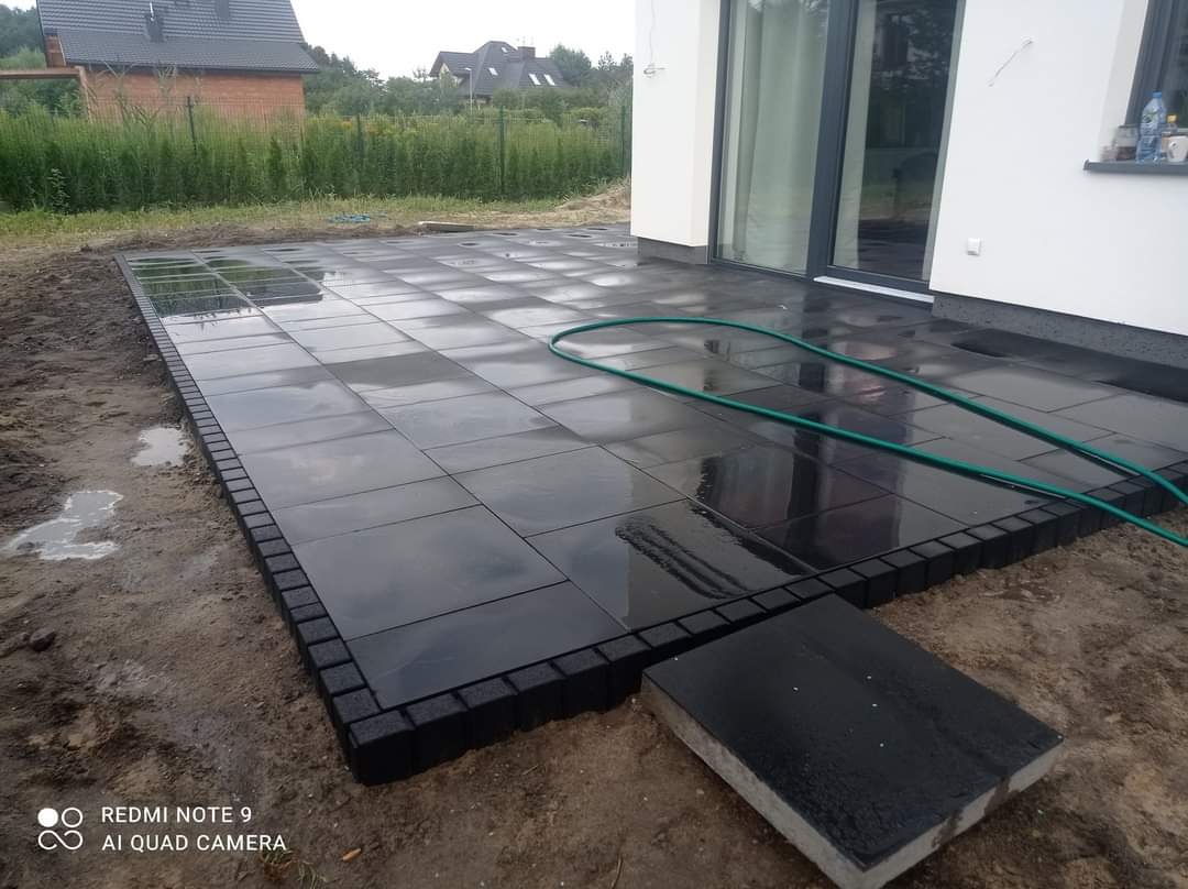 Świeżo ułożony, mokry taras z czarnej kostki brukowej z obrzeżem, widoczny wąż ogrodowy i fragment budynku w tle.