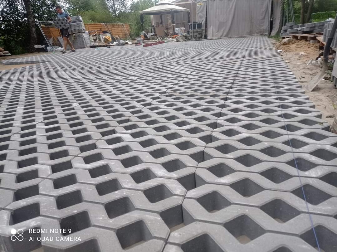 Ułożona nawierzchnia z ażurowych betonowych płyt w kształcie fal, widoczne niebieskie sznurki wyznaczające linię, w tle osoba przenosi kolejne elementy, teren budowy i zadaszenie.